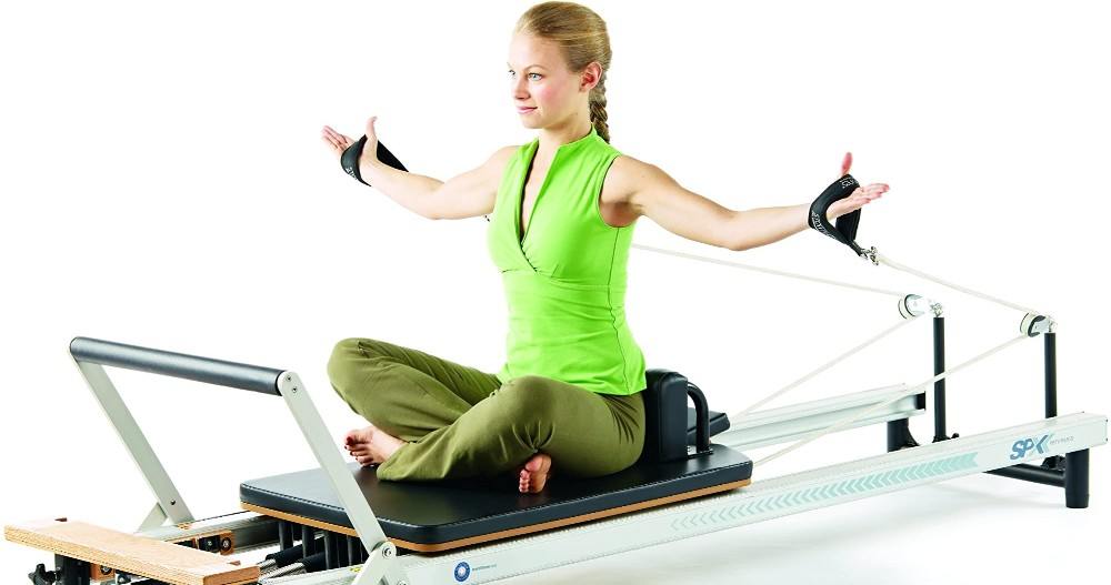 Woman using the Merrithew at Home SPX Reformer, sitting on the reformer facing foot bar with hands in short straps with arms out wide