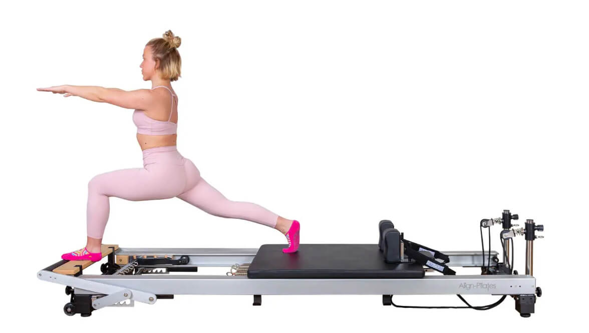 Woman using the Align Pilates Reformer, facing the front standing on foot platform and one foot back on the carriage.